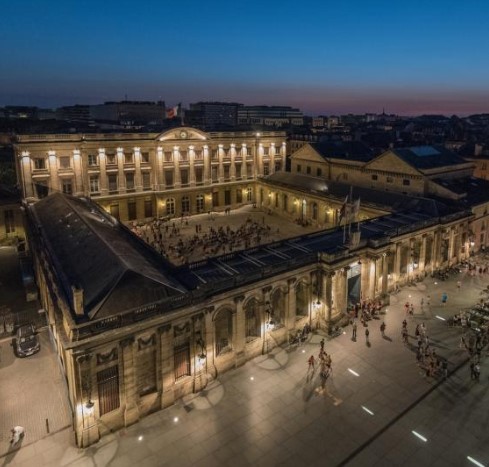 Vue nocturne de la Place de la Bourse à Bordeaux avec ses bâtiments historiques illuminés en jaune doré et des personnes circulant dans la place pavée.