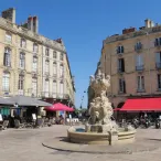 Place de la Bourse à Bordeaux avec fontaine ornementale, bâtiments historiques en pierre de taille, restaurants avec auvents rouges, et passants sous un ciel bleu.