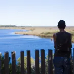 Une personne de dos observe un paysage avec un plan d'eau bleu et des marais salants, séparée par une barrière en bois, dans la région de Bordeaux.