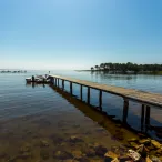 Un long ponton en bois s'étend dans une baie calme avec un petit bateau amarré, des arbres en arrière-plan et un ciel bleu clair, paysage fluvial typique de la région de Bordeaux.