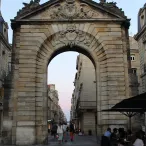 Porte de la Grosse Cloche à Bordeaux, imposant arc de pierre de style Renaissance avec bas-reliefs, donnant sur une rue pavée avec façades haussmanniennes et terrasse de café en premier plan.