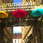 Entrée de la Promenade Sainte-Catherine à Bordeaux avec trois parapluies colorés (jaune, rouge, turquoise) suspendus au-dessus de la rue, et des centaines de parapluies multicolores visibles en arrière-plan près du port.