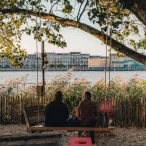 Deux personnes assises sur un banc sous un grand arbre au bord de la Garonne à Bordeaux, avec vue sur les façades historiques et les bâtiments modernes de la ville de l'autre côté du fleuve.