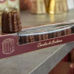 Canelé de Bordeaux dans sa boîte d'emballage traditionnelle marron et or, posé sur une surface grise avec d'autres canelés en arrière-plan