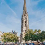 La Basilique Saint-Michel de Bordeaux avec sa flèche gothique élancée dominant une place animée remplie de terrasses de cafés et de visiteurs sous un ciel bleu.
