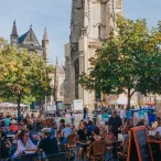 Vue d'une terrasse animée sur une place de Bordeaux avec la basilique Saint-Michel en arrière-plan, sous un ciel bleu dégagé.