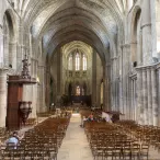 Intérieur de la Cathédrale Saint-André de Bordeaux montrant la nef centrale avec ses voûtes gothiques, ses colonnes en pierre, ses bancs en bois et l'autel illuminé au loin.