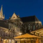 Vue nocturne de la Basilique Saint-Michel de Bordeaux avec ses flèches gothiques illuminées, entourée de bâtiments historiques et de terrasses ombragées en premier plan.