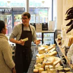 Un fromager sourit en montrant du fromage à une cliente dans sa boutique bordelaise. Le comptoir est rempli de fromages français variés et de produits gastronomiques.