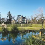 Château viticole bordelais entouré de vignobles, avec un étang de réflexion au premier plan et des arbres en arrière-plan sous un ciel dégagé.