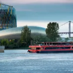 Un bateau de croisière rouge navigue sur la Garonne devant la Cité du Vin et le pont de Pierre à Bordeaux, avec l'architecture moderne du musée en arrière-plan