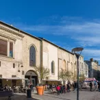 Façade du Cinéma Utopia à Bordeaux, bâtiment historique en pierre avec arcade, terrasse avec parasols, place publique animée avec visiteurs et bâtiments haussmanniens en arrière-plan.