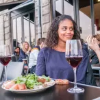 Deux femmes dégustent du vin rouge et un plat de fruits de mer en terrasse à Bordeaux, entourées d'autres clients dans un restaurant urbain.