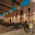 Soirée animée dans la cour intérieure de Darwin écosystème à Bordeaux, avec des visiteurs assis autour de tables, sous une structure métallique couverte et des arcades en pierre illuminées au coucher du soleil.