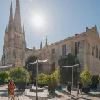 Place de la Cathédrale à Bordeaux avec la Basilique Saint-Michel en arrière-plan, des cyclistes et des piétons circulant dans l'espace public aménagé avec des voiles d'ombrage et de la végétation.