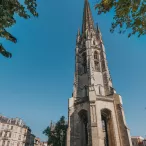 La Flèche Saint-Michel de Bordeaux, église gothique avec son clocher élancé, photographiée depuis une place entourée de bâtiments haussmanniens sous un ciel bleu dégagé.