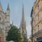 Vue de la flèche Saint-Michel de Bordeaux, église gothique avec sa tour pointue caractéristique, encadrée par des bâtiments haussmanniens et une boutique JULO en bas à droite.