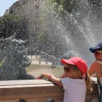 Deux enfants se rafraîchissent dans les jets d'eau d'une fontaine à Bordeaux par une journée ensoleillée, avec la place et ses bâtiments historiques en arrière-plan.