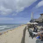 Plage avec baigneurs, terrasse de restaurant avec parasols, roue panoramique en arrière-plan et bâtiments côtiers à Bordeaux