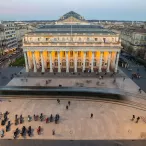 Vue aérienne de la Place de la Comédie à Bordeaux avec le Grand-Théâtre illuminé au centre, entouré de bâtiments haussmanniens et de personnes se promenant sur la place.