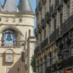 La Porte Cailhau à Bordeaux, tour médiévale en pierre avec trois tourelles coniques, horloge et cloche, entourée d'immeubles haussmanniens dans une rue historique.