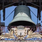 La Grosse Cloche de Bordeaux, une imposante cloche en bronze suspendue dans un beffroi en bois, encadrée par une ornementale grille en fer forgé doré aux motifs baroques et un blason sculpté.