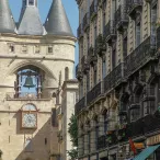 La Porte Cailhau, monument médiéval avec trois tours coniques et une horloge, entourée d'immeubles haussmanniens dans une rue de Bordeaux