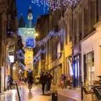 Vue nocturne d'une rue piétonne de Bordeaux illuminée avec la Grosse Cloche en arrière-plan, décorée de guirlandes bleues et blanches, avec des commerces et des passants.