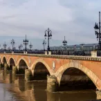 Un train moderne traverse le pont de Pierre de Bordeaux, un pont en brique rouge avec des arches caractéristiques enjambant la Garonne, avec la skyline de la ville en arrière-plan.