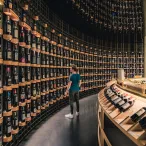 Intérieur d'une cave à vin moderne de Bordeaux avec des murs couverts de bouteilles de vin disposées sur des étagères en bois, un visiteur en tee-shirt bleu observant la collection, et des présentoirs de dégustation au premier plan.