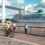 Vue du port de Bordeaux avec un grand paquebot de croisière amarré près du Pont de Pierre, des promeneurs et cyclistes sur la promenade fluviale.