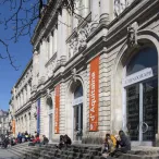 Façade du Musée d'Aquitaine à Bordeaux avec bannières oranges et bleues, visiteurs sur les marches, architecture néoclassique avec colonnes et ornements.