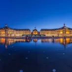 La Place de la Bourse de Bordeaux illuminée à la nuit tombante, avec ses bâtiments classiques se reflétant dans le miroir d'eau, un tramway bleu passant devant, et des personnes marchant sur la surface mouillée.