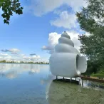 Installation artistique blanche en forme de coquillage au bord de l'eau à Bordeaux, avec la ville en arrière-plan et des arbres verts.