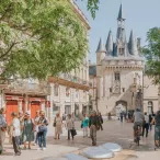 Place de la Grosse Cloche à Bordeaux avec la tour médiévale de la Grosse Cloche, des bâtiments historiques aux façades rouges, des passants, des cyclistes et des arbres verdoyants sous un ciel dégagé.
