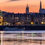 Vue crépusculaire de Bordeaux avec la Porte Cailhau illuminée au premier plan, les façades classiques du quai, les flèches de la cathédrale Saint-André en arrière-plan, et la Garonne en avant-plan avec un bateau de croisière.