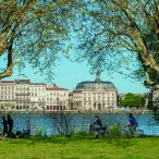 Vue de la Garonne à Bordeaux avec des bâtiments historiques en arrière-plan, des arbres centenaires au premier plan, et des personnes pratiquant des loisirs au bord de l'eau.