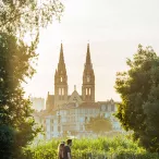 Deux personnes marchent sur un chemin bordé d'arbres avec la Basilique Saint-Michel de Bordeaux visible en arrière-plan au coucher du soleil.