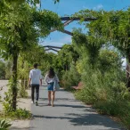 Deux personnes marchent sur un sentier verdoyant du Parc aux Angéliques à Bordeaux, encadrées par une végétation luxuriante et des structures métalliques arquées.