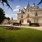 Château de style Belle Époque avec tours pointues et façade en pierre beige, entouré d'un parc paysager avec pelouse verte et parterre de fleurs colorées, situé dans la région de Bordeaux.