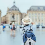 Touriste de dos portant un chapeau blanc et un sac à dos bleu, contemplant la Place de la Bourse et ses bâtiments historiques à Bordeaux