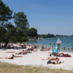 Plage aménagée au bord d'un lac avec de nombreux baigneurs, des voiliers, une zone de baignade délimitée et des arbres en arrière-plan sous un ciel dégagé.