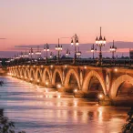 Le Pont de Pierre de Bordeaux illuminé au coucher du soleil, avec ses arches en pierre et ses réverbères allumés se reflétant dans l'eau de la Garonne