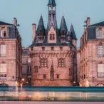 Château de la Malartrie à Bordeaux avec ses tours pointues en pierre rose illuminées au crépuscule, flanqué de bâtiments mansardés et reflété dans l'eau