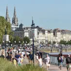 Vue panoramique de la place de la Bourse à Bordeaux avec ses façades classiques du XVIIIe siècle, la basilique Saint-Michel en arrière-plan, la Garonne et les quais animés par de nombreux visiteurs par beau temps.