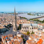 Vue aérienne de Bordeaux montrant la Basilique Saint-Michel avec sa flèche caractéristique, la Garonne avec ses ponts, et l'urbanisation dense de la ville historique aux toits orangés.