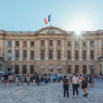 La Cour du Parlement de Bordeaux avec sa façade classique en pierre, le drapeau français flottant au-dessus, et des visiteurs rassemblés sur la place pavée sous un ciel dégagé.