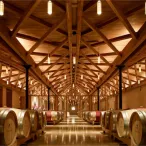 Intérieur d'un chai de Bordeaux avec des rangées de fûts de chêne alignés symétriquement sous une structure architecturale en bois aux poutres apparentes, éclairé par des luminaires modernes.
