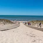 Vue panoramique d'une plage de sable fin avec une promenade en bois central, des dunes végétalisées de chaque côté, et de nombreux baigneurs dans l'eau turquoise sous un ciel dégagé.
