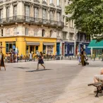 Une place publique à Bordeaux avec des bâtiments haussmanniens, un commerce jaune vif au rez-de-chaussée, des terrasses de café avec parasols, des arbres verts, et des passants se détendant sur des bancs.
